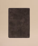 The Suede Passport Holder, a travel essential in dark brown with a soft, textured finish and subtle embossed text near the bottom, is centered on a light beige background.