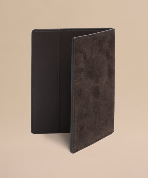 A Suede Passport Holder in brown is partially open on a beige background, displaying its smooth leather interior and textured suede exterior—perfect for your travel essentials.