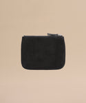 The Suede Pouch is a small black suede accessory with a zippered closure and minimalist design, perfect for use as a compact pouch or passport holder, pictured on a plain beige background.