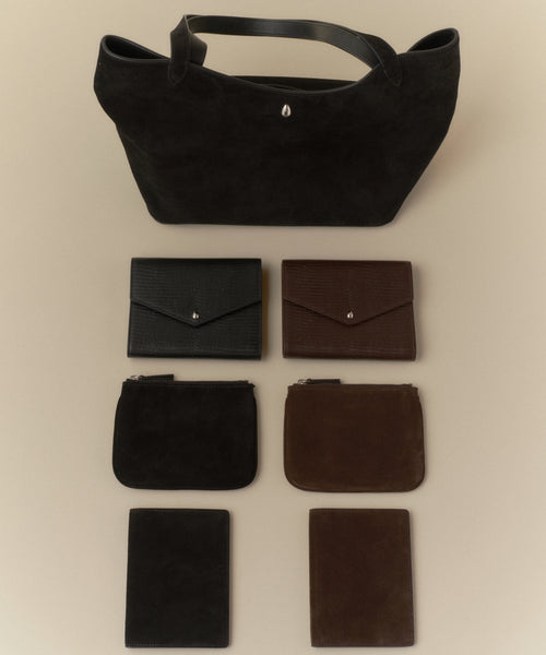 A black tote bag sits above three rows: two wallets (black and brown), a Suede Pouch, and two matching rectangular card holders, all arranged neatly on a beige surface.