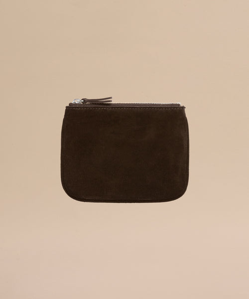 The Suede Pouch is a small, square, dark brown suede pouch with a silver zipper, shown against a plain beige background.