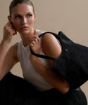 A woman with pulled-back hair wears a sleeveless white top, gold teardrop stud earrings, and carries the Suede Ryder Bag over her shoulder as she gazes thoughtfully to the side against a neutral background.