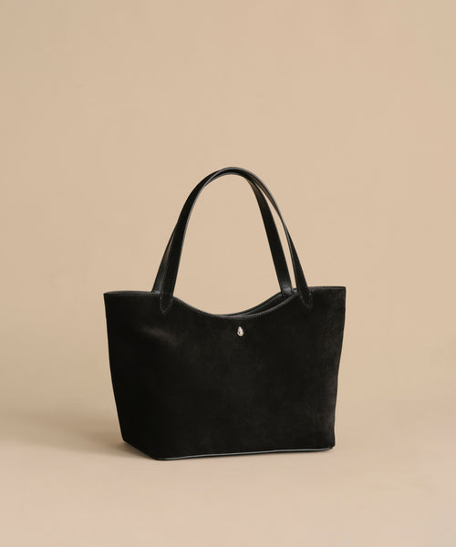 The Suede Ryder Bag in black suede with leather handles is showcased on a beige background. This stylish shoulder bag features a minimalist design, magnetic closure, and a metallic accent on the front.