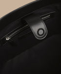 Close-up of the inside of the Suede Ryder Bag, featuring a zippered pocket and a leather tab with a round magnetic closure, set against a beige background.