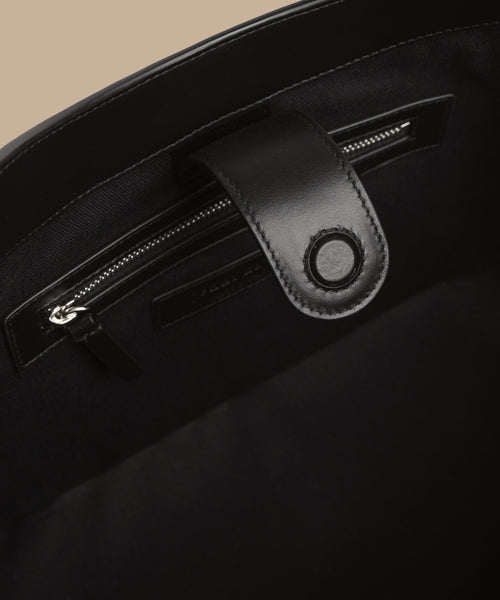 Close-up of the inside of the Suede Ryder Bag, featuring a zippered pocket and a leather tab with a round magnetic closure, set against a beige background.