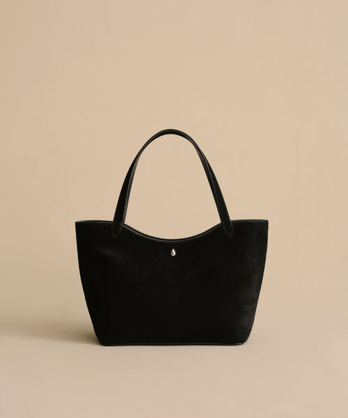 A black Suede Ryder Bag tote with two handles, shown on a beige background. This minimalist bag has a subtle silver button and a magnetic top-center closure for convenience.