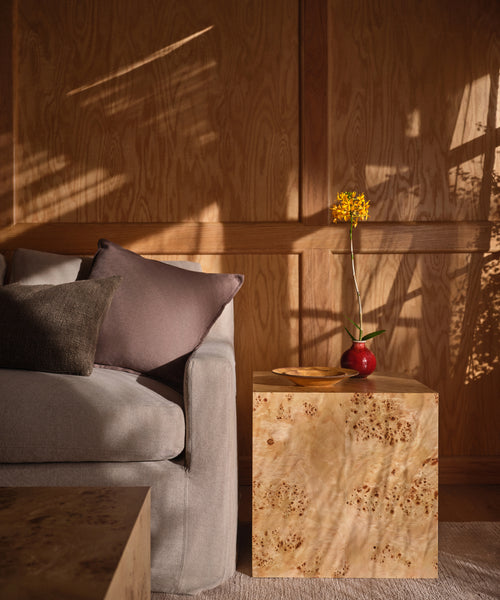 A cozy living room corner with a light gray sofa, two pillows, and the Sur Side Table topped with a small red vase and yellow flower, bathed in warm sunlight and soft shadows for a vintage furniture vibe.