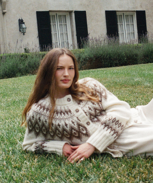 A woman with long brown hair reclines on green grass, wearing the Thatcher Crewneck Cardigan—a cream-colored sweater with a brown geometric pattern. Behind her is a beige building with black shutters and lavender bushes.