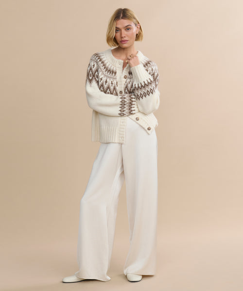 A woman with a blonde bob stands against a beige background, wearing the Thatcher Crewneck Cardigan, wide-leg white pants, and white shoes. She poses with one hand touching her face.