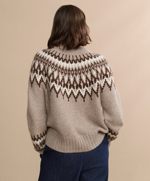 A person with shoulder-length brown hair is facing away, wearing the beige Thatcher Crewneck sweater featuring a brown and white geometric pattern and dark blue pants, set against a plain beige background.
