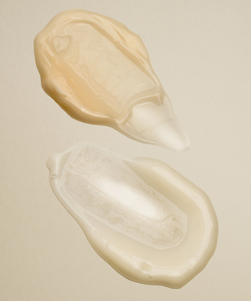 Two translucent blobs of liquid, one pale peach and one clear, are spread on a smooth beige surface—evoking a luxe skincare routine with The Serums in organic, fluid shapes.