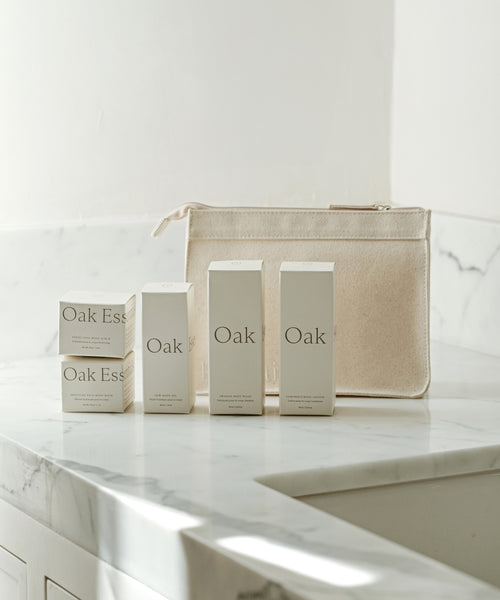 Four cream-colored TSA-approved minis from The Travel Body Routine by Oak Ess are set on a marble countertop beside a matching pouch in a softly lit, minimalist bathroom—your perfect travel skincare essentials.