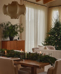 A cozy dining room decorated for Christmas, featuring a handcrafted Trestle Dining Table adorned with greenery and candles, a modern light fixture, a sideboard with a potted plant, and a Christmas tree with ornaments by sheer curtained windows.