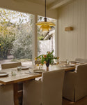 A cozy dining room features the Trestle Dining Table set for six, beige upholstered chairs, a yellow pendant light, and large windows overlooking a sunny garden. Fresh flowers on the table add to the inviting atmosphere.