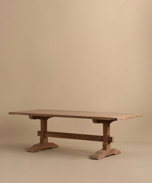 The Trestle Dining Table features a handcrafted, smooth top and sturdy trestle legs. Its simple, modern design showcases natural wood grain for timeless appeal, displayed against a plain beige background.