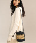 A woman in a cream sweater and beige skirt carries the Tropez Bucket Bag—a handcrafted black and tan cane accessory with a long shoulder strap—standing against a Tuscany-inspired neutral background.