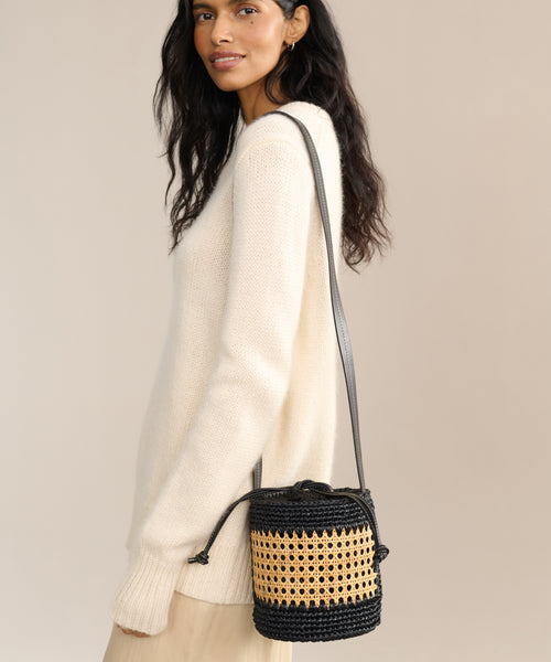 A woman in a cream sweater and beige skirt carries the Tropez Bucket Bag—a handcrafted black and tan cane accessory with a long shoulder strap—standing against a Tuscany-inspired neutral background.