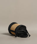 The small Tropez Bucket Bag, crafted with black and tan rattan-style weaving, black leather strap, and a black drawstring closure, rests on its side against a plain beige background.