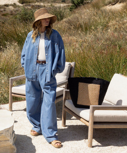 A person wearing a wide-brimmed hat, oversized denim shirt, wide-leg jeans, and sandals stands outdoors by cushioned chairs with the Tropez Tote Bag, surrounded by tall grass and shrubs.
