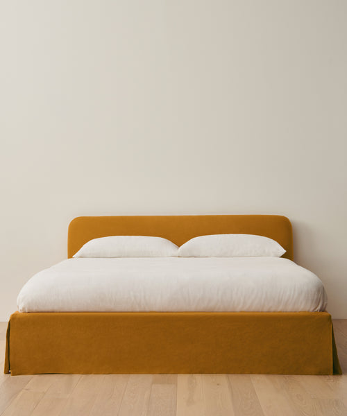 The Cove Bed showcases a mustard linen upholstered headboard and frame, accented with crisp white bedding and two pillows, set on a light wood floor beside a plain beige wall.