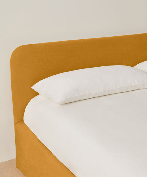 The Cove Bed boasts a mustard linen upholstered headboard and frame, dressed in crisp white bedding with a single white pillow, set against a light-colored wall.