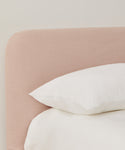 A close-up of a Cove Bed featuring a light pink linen upholstered headboard in Belgian Libeco Linen, accented with a white pillow and bedding, set against a plain light-colored wall.