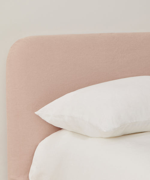 A close-up of a Cove Bed featuring a light pink linen upholstered headboard in Belgian Libeco Linen, accented with a white pillow and bedding, set against a plain light-colored wall.