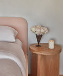 A wooden bedside table with a white candle and metal vase of white flowers stands by the Cove Bed, featuring a linen upholstered headboard and Belgian Libeco linen bedding. The wall behind is light gray.