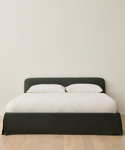 The Cove Bed, featuring a Belgian linen headboard, is dressed with two white pillows and a white duvet and placed on a light wooden floor against a plain, light-colored wall.