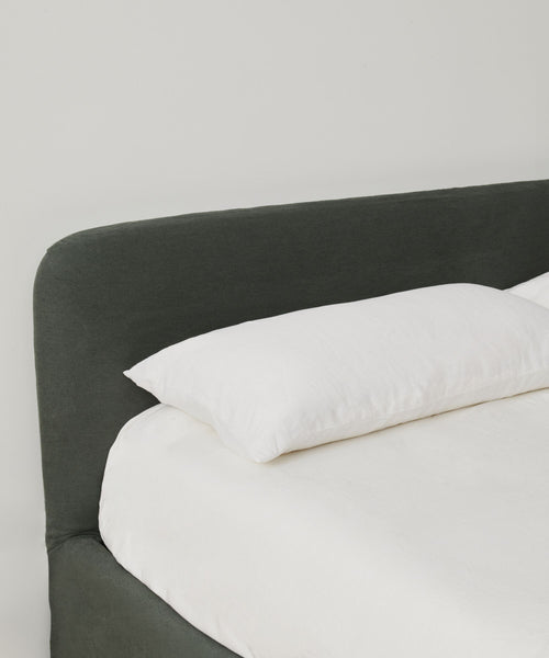 The Cove Bed features a dark upholstered headboard and a single white pillow on crisp white sheets, all set against a plain white wall.