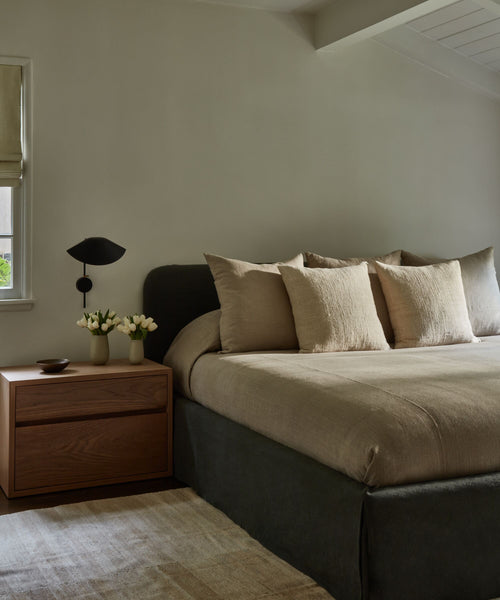 A modern bedroom showcases the Cove Bed with a Belgian linen beige headboard, four matching pillows, a wooden nightstand holding two white tulip vases and a bowl, plus a black wall sconce. Neutral walls and a soft rug create a serene atmosphere.