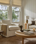 Bright living room with large windows, a white Harbor Chair, an ivory linen sofa, beige throw blanket, wooden coffee table with books, vase of flowers, candle, and side table with lamp. Trees are visible outside the windows.
