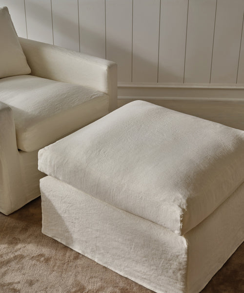 An ivory linen armchair and the handmade in Los Angeles Harbor Ottoman sit atop a beige carpet against a white paneled wall in a softly lit room.