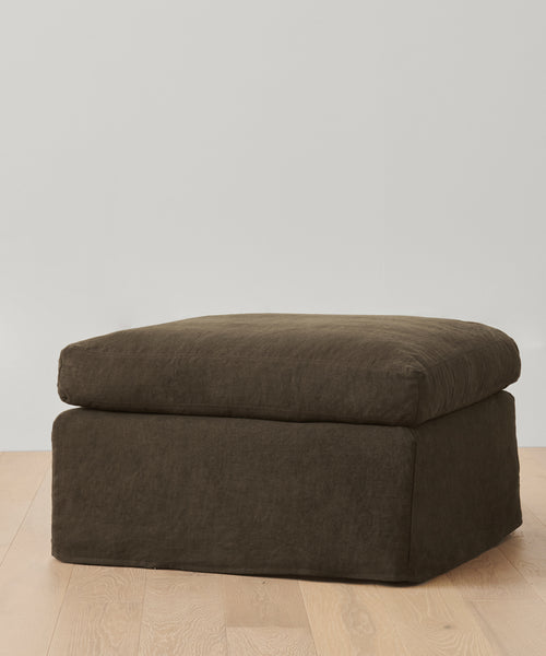 A dark brown Harbor Ottoman from Jenni Kayne Furniture, featuring a cushioned top, sits on a light wooden floor against a plain white wall for an elegant touch.