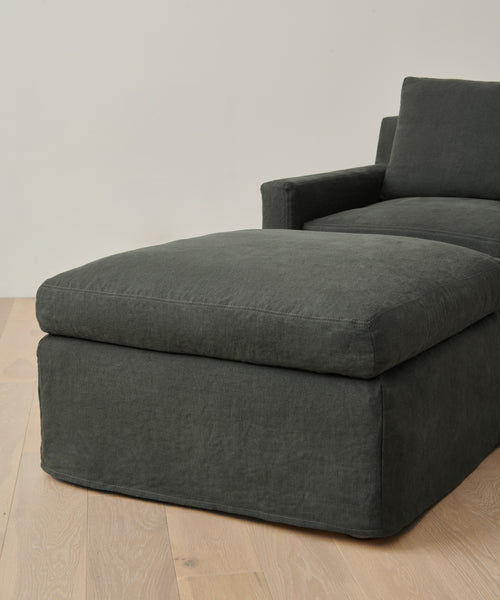 A dark grey upholstered Harbor Ottoman from Jenni Kayne Furniture, with a matching cushion, rests on a light wooden floor against a plain, light-toned wall.