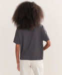 A person with voluminous curly hair is seen from the back, wearing a loose-fitting Vintage Pocket Tee in charcoal and light pants against a plain background for a relaxed, casual look.