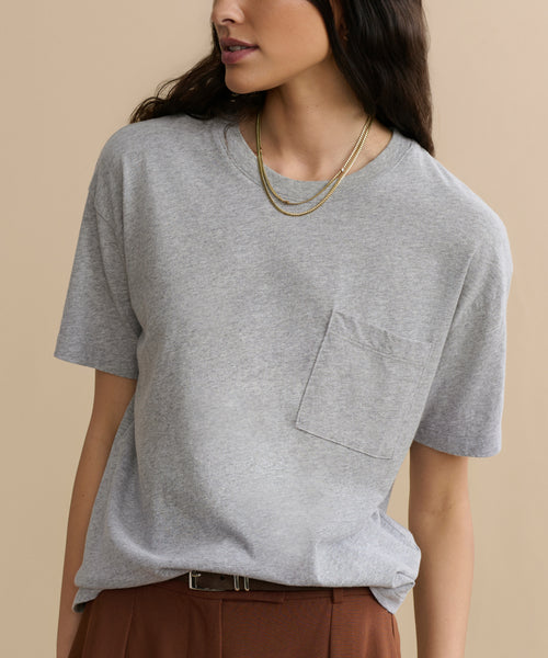 A woman wearing a loose-fitting Vintage Pocket Tee in heather grey, paired with brown pants and layered gold necklaces, stands against a beige background.
