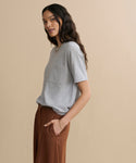 A woman with long dark hair stands sideways against a beige background, wearing the Vintage Pocket Tee in Heather Grey and high-waisted brown pants, hands in her pockets.