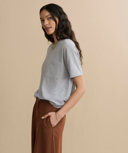 A woman with long dark hair stands sideways against a beige background, wearing the Vintage Pocket Tee in Heather Grey and high-waisted brown pants, hands in her pockets.