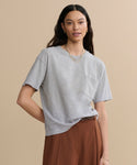 A woman with long dark hair wears the Vintage Pocket Tee in heather grey with brown high-waisted pants, accessorized with gold hoop earrings and layered necklaces, standing against a beige background.