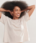 A woman with voluminous curly hair smiles with her arms raised, wearing the Vintage Pocket Tee—a light beige, short-sleeve top with a chest pocket for a casual look.