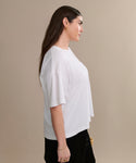 A woman with long brown hair stands in profile against a beige background, wearing the Vintage Pocket Tee in white, paired with black pants and gold jewelry.