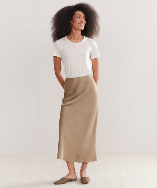 A woman with curly hair smiles while wearing a white short-sleeve t-shirt tucked into the Sawyer Slip Skirt and brown flats, standing against a plain light background.