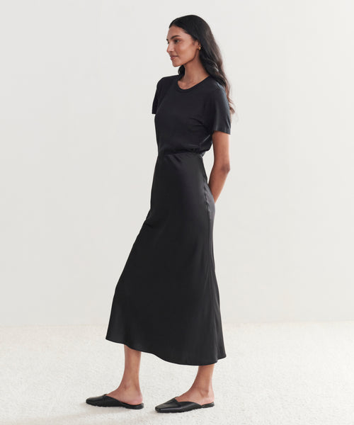 A woman stands sideways, wearing a short-sleeve black t-shirt tucked into the Sawyer Slip Skirt and black slip-on shoes, posing against a simple light background.