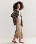 A woman with voluminous curly hair wears a brown cardigan over a white top, the tan Sawyer Slip Skirt, and brown slip-on shoes, smiling as she stands sideways against a light neutral background.