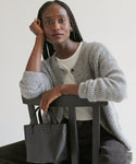 A woman with long braided hair sits on a chair, resting her head on her hand. She wears the Hayes Crewneck Cardigan over a white shirt and holds a black handbag, set against a plain, light-colored background.