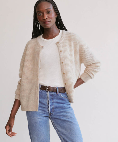 A woman with long braided hair wears the Hayes Crewneck Cardigan in cream over a white shirt, blue jeans, and a brown belt, standing against a plain light background.
