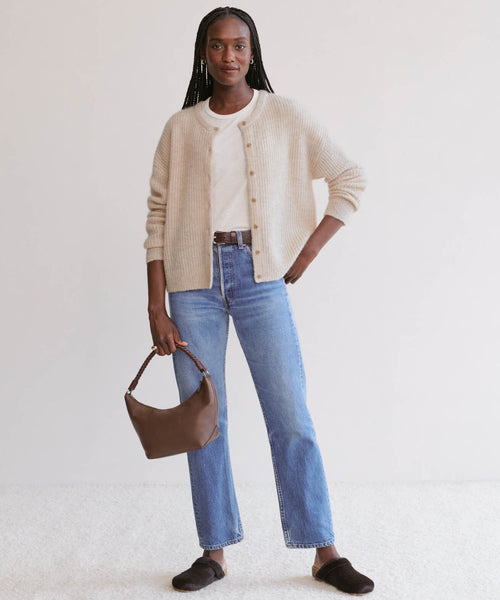 A woman with long braids smiles in the Hayes Crewneck Cardigan, styled over a white shirt, blue jeans, and dark slippers. She holds a brown handbag and poses against a light background with one hand on her hip.