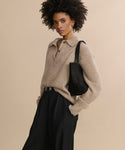 A woman with curly hair wears a beige collared sweater and black Wool Jones Trouser, standing against a beige background. She holds a black shoulder bag and poses confidently, highlighting the versatile style of the Wool Jones Trouser.