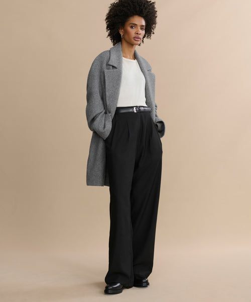 A woman with short curly hair and a confident expression stands against a beige background wearing the Wool Jones Trouser, paired with a gray coat and white top. Her hands are in her pockets, showcasing this modern, versatile pant inspired by Italian suiting.
